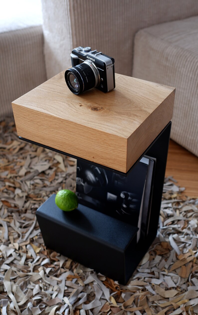 Op de afbeelding: Een zwarte metalen bijzettafel met een houten blad. De tafel heeft een plank voor opslag en staat op een tapijt gemaakt van leren stroken.