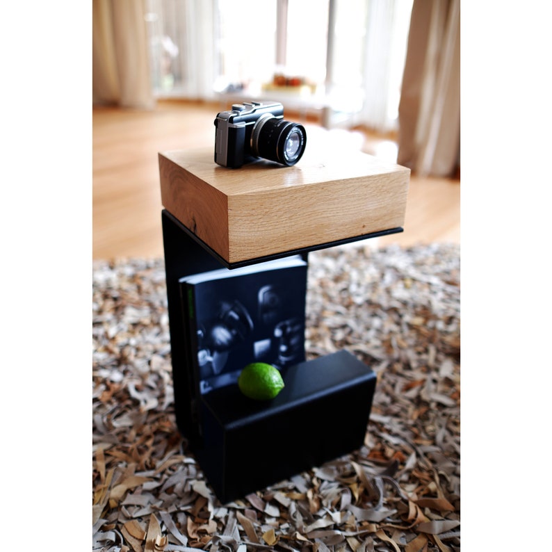 Op de afbeelding: Een zwarte metalen bijzettafel met een houten blad. De tafel heeft een plank onder het blad. Een camera en een limoen liggen op het blad van de tafel.
