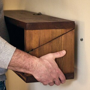 May include: A person installing a wooden wall shelf with a hinged door. The shelf is made of dark brown wood and has a simple design. The shelf is being attached to a light beige wall with screws.