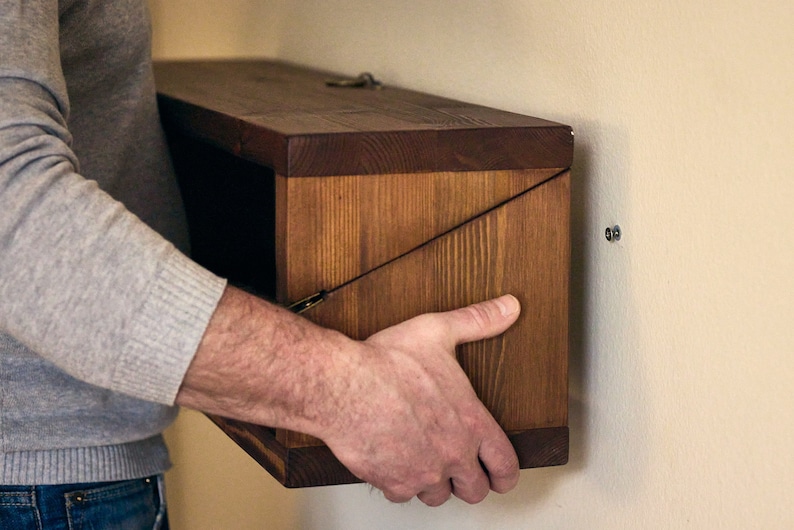 May include: A person installing a wooden wall shelf with a hinged door. The shelf is made of dark brown wood and has a simple design. The shelf is being attached to a light beige wall with screws.