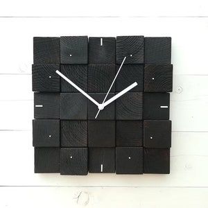Puede incluir: Un reloj de madera negro con manecillas blancas y un dise&ntilde;o minimalista. El reloj est&aacute; hecho de bloques de madera cuadrados individuales.
