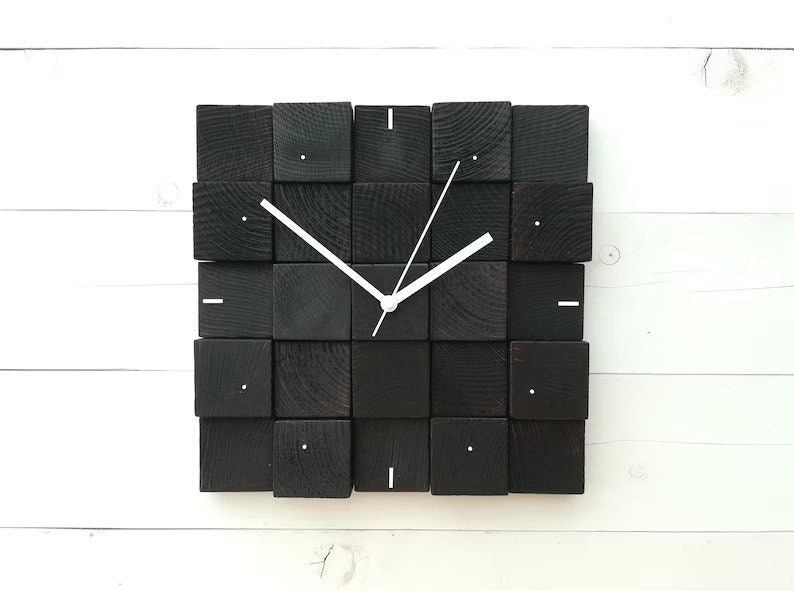 Puede incluir: Un reloj de madera negro con manecillas blancas y un dise&ntilde;o minimalista. El reloj est&aacute; hecho de bloques de madera cuadrados individuales.