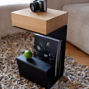 Op de afbeelding: Een zwarte metalen bijzettafel met een houten blad. De tafel heeft een plank voor tijdschriften of boeken en een kleine lade. Een vintage camera staat op het tafelblad.
