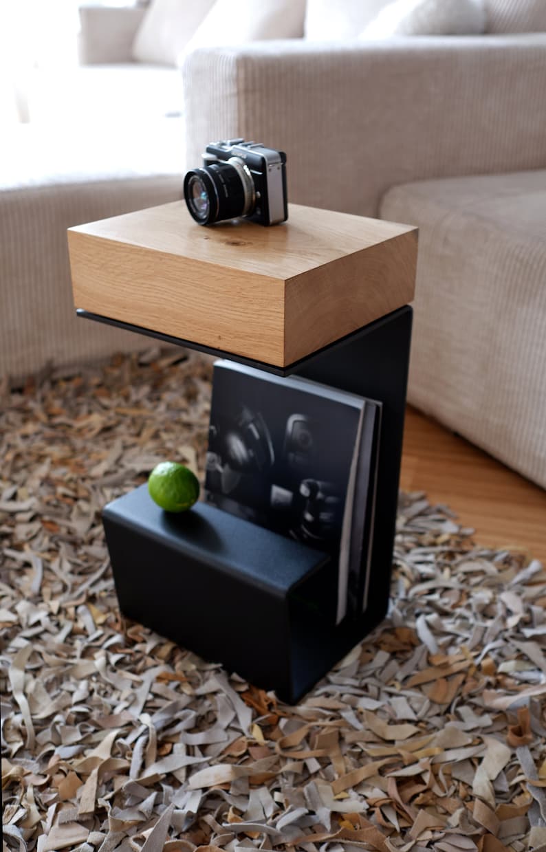 Op de afbeelding: Een zwarte metalen bijzettafel met een houten blad. De tafel heeft een plank voor tijdschriften of boeken en een kleine lade. Een vintage camera staat op het tafelblad.