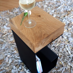 Op de afbeelding: Een houten bijzettafel met een zwart metalen frame en een plank. De tafel heeft een natuurlijke houten afwerking en is bedekt met een glas witte wijn en een bloem.