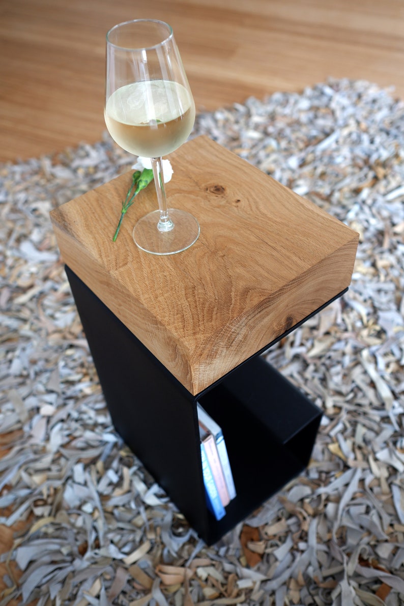 Op de afbeelding: Een houten bijzettafel met een zwart metalen frame en een plank. De tafel heeft een natuurlijke houten afwerking en is bedekt met een glas witte wijn en een bloem.