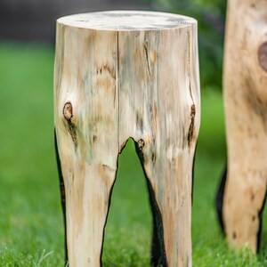 May include: A wooden stool made from a tree trunk with a natural wood finish. The stool has a round top and three legs. The stool is sitting on a green grassy surface.