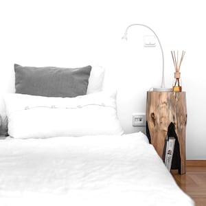 May include: A white bed with a gray pillow and a white pillow with buttons. A wooden nightstand with a lamp and a diffuser is next to the bed.