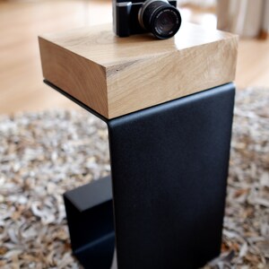 Op de afbeelding: Een zwarte metalen bijzettafel met een houten blad. De tafel heeft een eenvoudig, modern ontwerp en is perfect voor een kleine ruimte. Een zwarte camera staat op het houten blad.