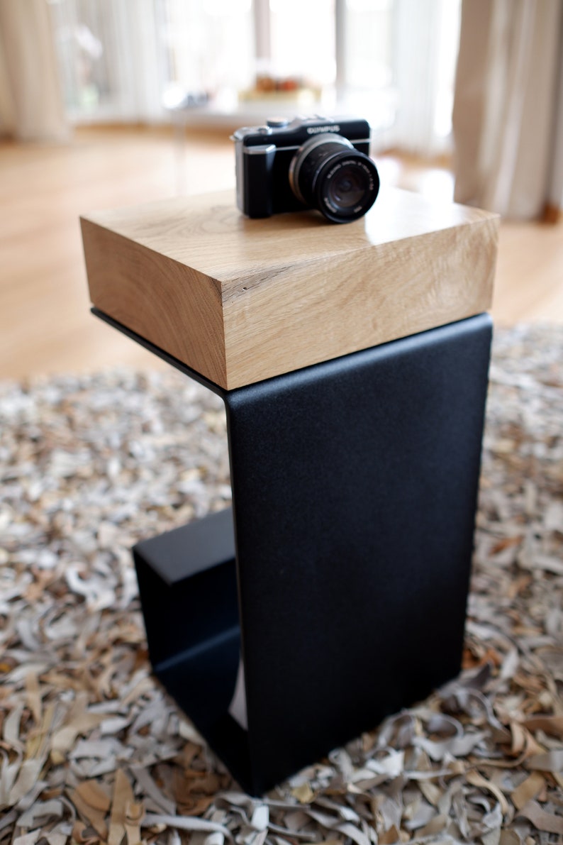 Op de afbeelding: Een zwarte metalen bijzettafel met een houten blad. De tafel heeft een eenvoudig, modern ontwerp en is perfect voor een kleine ruimte. Een zwarte camera staat op het houten blad.