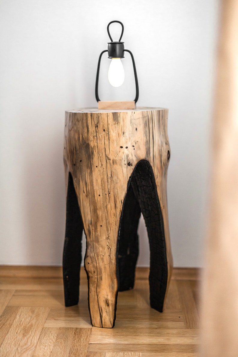May include: A rustic wooden side table with black painted legs. The table has a natural wood grain and is topped with a black metal lamp with a light bulb.