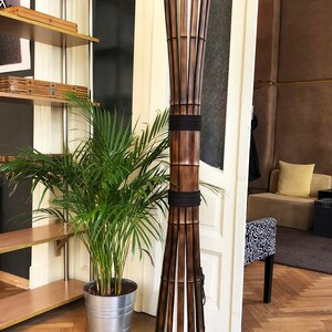 Op de afbeelding: Een hoge, bruine bamboe vloerlamp met een witte stoffen kap. De lamp is vastgebonden met zwart touw en heeft een zwart snoer.
