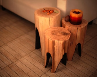 Op de afbeelding: Drie houten boomstronk krukken van verschillende hoogtes. De krukken zijn lichtbruin met zichtbare houtnerf en donkere accenten. Eén kruk heeft een kaars met oranje, rode en bruine tinten. Decoratieve items staan op de andere krukken.