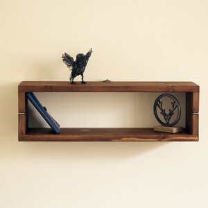 May include: A dark brown wooden shelf with a wire bird sculpture, a book, and a metal deer head sculpture on top. The shelf has a rectangular shape and is mounted on a white wall.