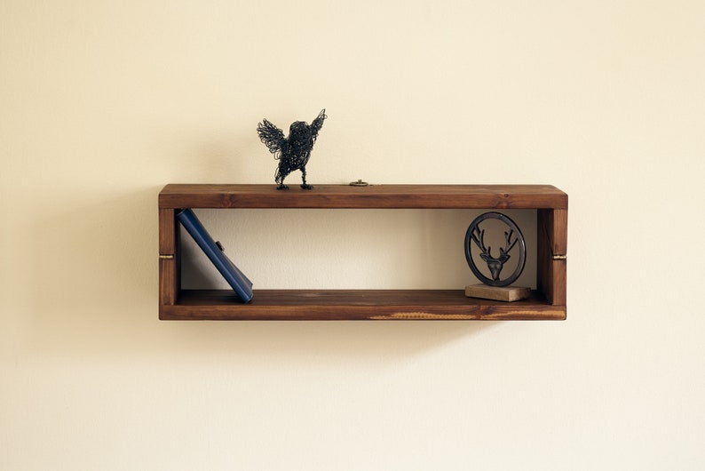 May include: A dark brown wooden shelf with a wire bird sculpture, a book, and a metal deer head sculpture on top. The shelf has a rectangular shape and is mounted on a white wall.