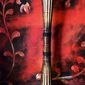 Op de afbeelding: Een hoge, slanke vloerlamp gemaakt van natuurlijke bamboe met een lichtbruine afwerking. De lamp heeft een uniek ontwerp met een brede basis en een smalle bovenkant. De lamp is aan en geeft een warm licht. De lamp staat voor een rode muur met een bloemenmotief.