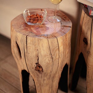 May include: Two wooden side tables made from tree trunks with natural bark and knots. The table on the left has a glass bowl with almonds on it and a glass of white wine. The table on the right is empty.