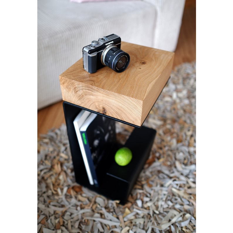Op de afbeelding: Een zwarte metalen bijzettafel met een houten blad. De tafel heeft een plank voor boeken en een kleine groene appel op de plank. Een zwarte camera ligt op het houten blad.
