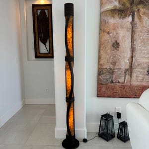 K&ouml;nnte beinhalten: Eine hohe, braune Stehlampe mit einem gewebten, zylindrischen Schirm. Der Schirm ist von innen beleuchtet und strahlt ein warmes Licht aus. Die Lampe steht auf einem hellen Fliesenboden neben zwei kleineren schwarzen Laternen.