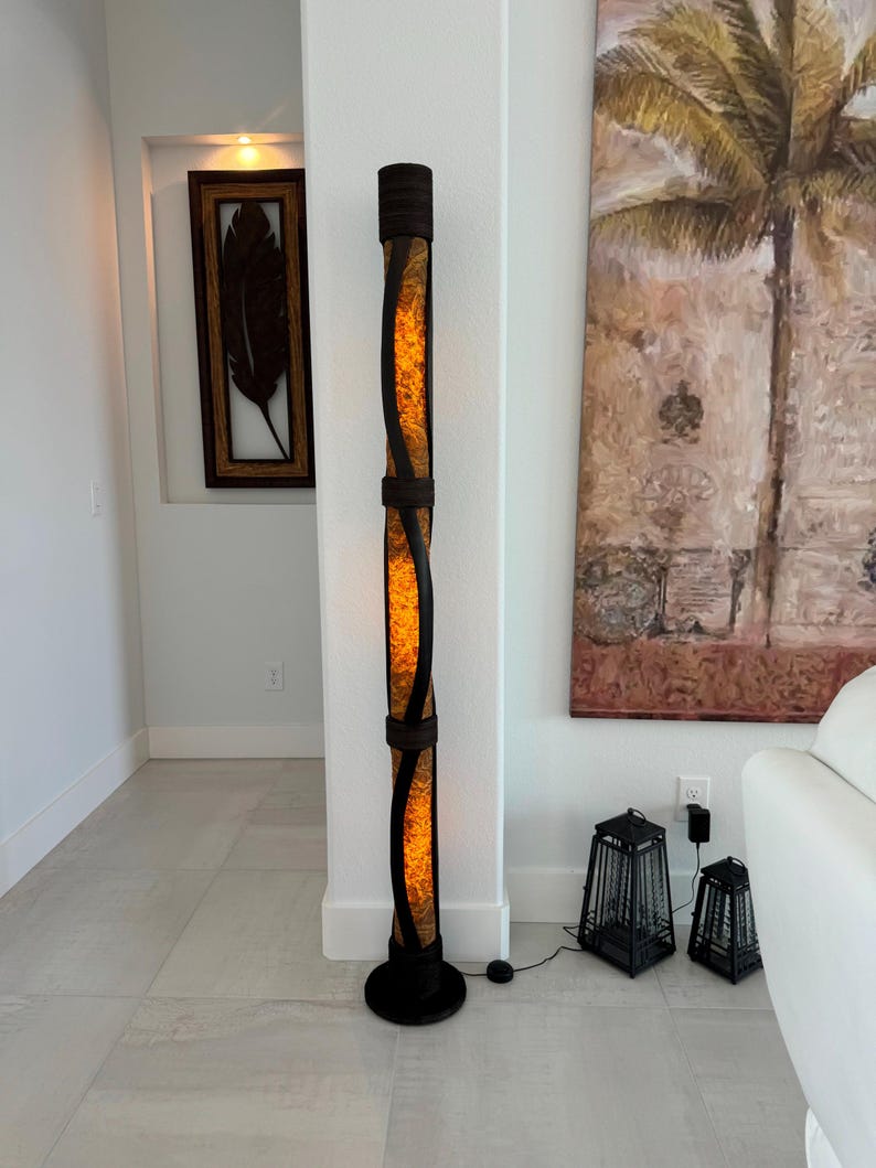 K&ouml;nnte beinhalten: Eine hohe, braune Stehlampe mit einem gewebten, zylindrischen Schirm. Der Schirm ist von innen beleuchtet und strahlt ein warmes Licht aus. Die Lampe steht auf einem hellen Fliesenboden neben zwei kleineren schwarzen Laternen.