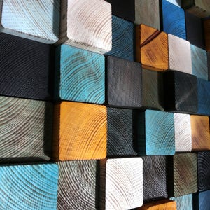 Puede incluir: Una pieza de arte de pared hecha de bloques de madera en varios colores, incluyendo azul, verde, negro, blanco y naranja. Los bloques est&aacute;n dispuestos en un patr&oacute;n geom&eacute;trico.