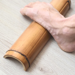 May include: A person's foot rolling over a natural brown bamboo roller on a light wood surface.