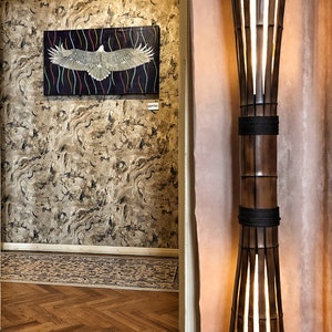 Op de afbeelding: Een hoge, houten vloerlamp met een lichtbruine afwerking en een witte, doorschijnende kap. De lamp is gemaakt van dunne, verticale latten hout en heeft een touwachtige band rond het midden. De lamp is aan en geeft een warm licht.