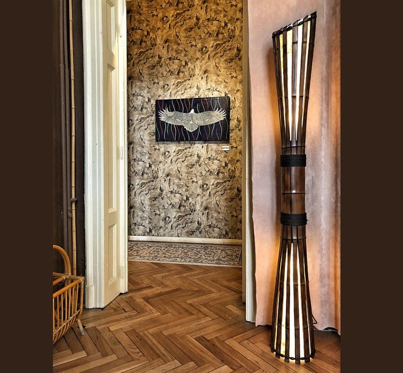 Op de afbeelding: Een hoge, slanke vloerlamp gemaakt van natuurlijke bamboe met een lichtbruine afwerking. De lamp heeft een brede, uitlopende voet en een smalle, cilindrische kap. De lamp is aan en geeft een warm licht.