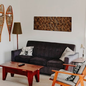 Puede incluir: Un interior de sala de estar con un sof&aacute; gris oscuro, una mesa de caf&eacute; de madera, un sill&oacute;n de madera, una l&aacute;mpara de pie y dos raquetas de nieve de madera colgadas en la pared. La pared detr&aacute;s del sof&aacute; tiene una obra de arte rectangular de mosaico de azulejos marrones.