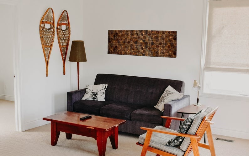 Puede incluir: Un interior de sala de estar con un sof&aacute; gris oscuro, una mesa de caf&eacute; de madera, un sill&oacute;n de madera, una l&aacute;mpara de pie y dos raquetas de nieve de madera colgadas en la pared. La pared detr&aacute;s del sof&aacute; tiene una obra de arte rectangular de mosaico de azulejos marrones.