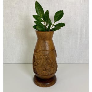 May include: A handcrafted wooden vase with a detailed carved design. The vase is filled with green leafy branches. The vase is brown and has a round base. The background is a white textured surface.