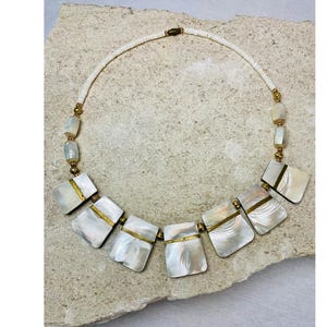 May include: A white shell necklace with square and rectangular shell pieces. The necklace has gold-colored metal accents and a white beaded strand. The necklace is displayed on a light-colored stone surface.