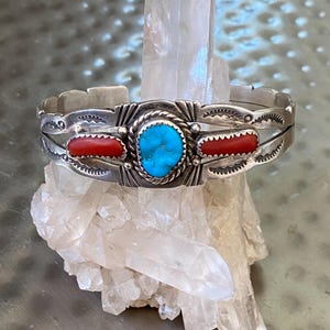 May include: Silver cuff bracelet with a central turquoise stone and two red coral accents. The bracelet has a detailed design with a rope-like border around the turquoise stone and decorative edges.