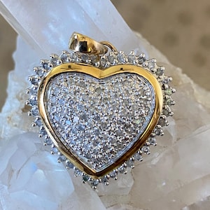 May include: A heart-shaped pendant with a gold border and a cluster of small, sparkling stones. The pendant has a gold bail for attaching to a chain. The outer edge is lined with smaller, clear stones, creating a halo effect. The pendant is likely made of gold and gemstones.