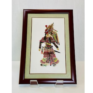 May include: A framed Chinese shadow puppet figure featuring intricate red, gold, and black patterns on a white background. The figure wears an elaborate headdress and traditional costume with detailed embroidery, encased in a dark wood frame.