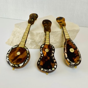 May include: Three miniature stringed instruments featuring brown tortoiseshell-patterned bodies with inlaid mother-of-pearl accents. Each piece includes a long neck with fret markings and a small circular sound hole, crafted with a glossy finish.