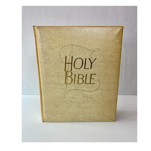 May include: A vintage Bible with a textured, light gold cover. The words "HOLY BIBLE" are printed in gold script on the front. The book is closed and standing upright on a white surface.