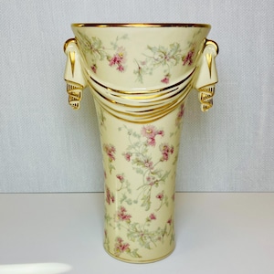 May include: A tall, cream-colored vase with a floral pattern of pink flowers and green leaves. The vase has gold trim and decorative handles. The design includes gold bands and draped accents, creating an elegant aesthetic.