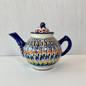 May include: A ceramic teapot with a lid, featuring a vibrant, intricate pattern in blue, orange, yellow, and brown. The spout and handle are a deep blue, and the lid has a floral design. A decorative piece for serving tea.