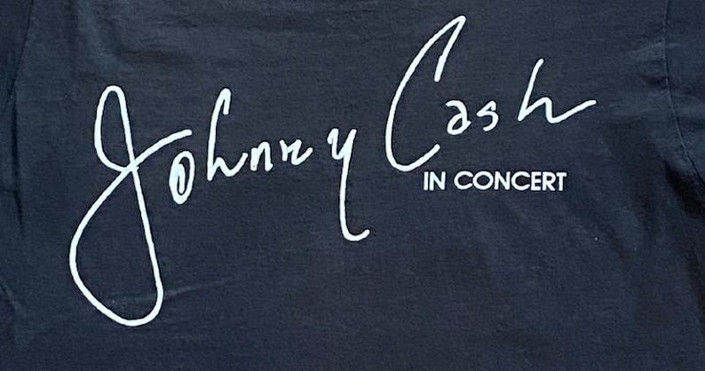 K&ouml;nnte beinhalten: Schwarzes T-Shirt mit dem wei&szlig;en Schriftzug "Johnny Cash IN CONCERT" in kursiver Schrift. Der Text sieht aus wie eine Unterschrift. Das T-Shirt besteht aus einem dunklen Stoff.