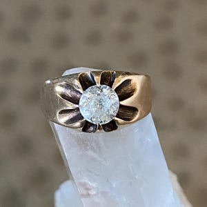 May include: A gold ring with a large, clear, round gemstone set in a flower-shaped design. The ring is displayed on a clear, crystalline surface, with a soft, patterned background.