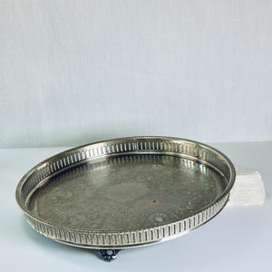 May include: An oval silver-toned serving tray featuring an intricate engraved floral pattern on the base and a raised gallery rim with a vertical slat design. The tray is supported by small, ornate feet and has a polished, reflective surface.