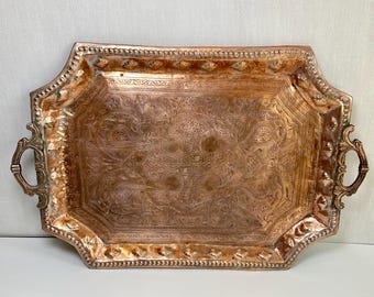 May include: A rectangular copper tray featuring intricate engraved floral and paisley patterns across the center. The tray has a raised, textured border with a repeating beaded design and two ornate, curved metal handles on the sides.