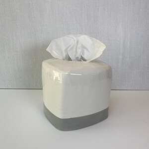 May include: A square, ceramic tissue box with a white upper section and a gray base. White tissues are visible, emerging from the top opening. The box has a glossy finish and a modern design, suitable for home or office use.
