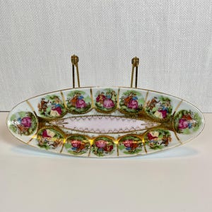 May include: Oval porcelain dish with a pink center and a decorative border. The dish features gold trim and painted scenes of people in period clothing. The dish is supported by two gold-colored metal stands.