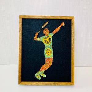 May include: A framed string art piece depicting a badminton player in action. The artwork features a player in a yellow and orange outfit, holding a racket, against a black background. The frame is made of wood.