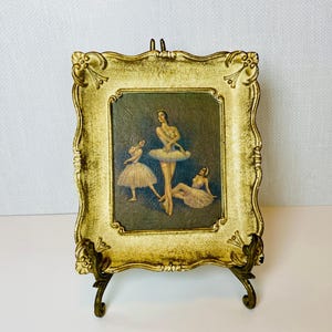 May include: An antique-style framed print featuring three ballerinas in a classical pose. The frame is ornate with a gold finish and a decorative stand. The artwork depicts the dancers in soft, muted colors, creating a vintage aesthetic.