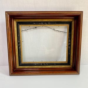 May include: A rectangular wooden picture frame with a dark brown finish. The frame has a layered design with gold and black accents. The inner frame is empty, with a wire for hanging.