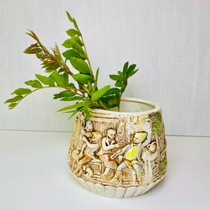 May include: Cream-colored ceramic vase with a detailed relief scene of figures in period clothing. The vase has a wide, slightly flared shape and is filled with green leafy branches. The relief depicts people in a tavern setting.