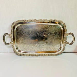 May include: A rectangular silver-colored serving tray with ornate handles and a decorative floral pattern. The tray has a scalloped edge and is likely made of metal. The handles are curved and add to the tray's elegant design.